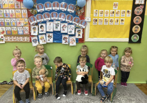 Dzieci pozują do zdjęcia przed tablicą z dekoracją. Chłopaki siedzą na krzesłach a dziewczynki stoją za nimi.