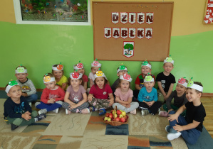 Cała grupa pozuje do zdjęcia na dywanie, pod tablicą z napisem „Dzień Jabłka” z opaskami jabłka na głowach. Przed nimi stoi koszyk pełen kolorowych jabłek.
