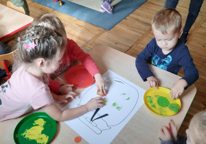 Dwie dziewczynki i dwóch chłopców przy stoliku odbijają umoczone w kolorowych farbach połówki jabłek na słoiku narysowanym na kartonie.