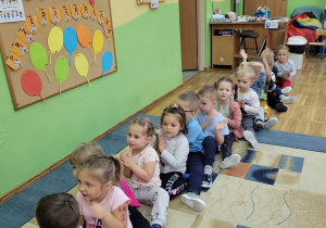 Dzieci siedzą na dywanie jeden za drugim śpiewając piosenkę i klaszczą w dłonie.