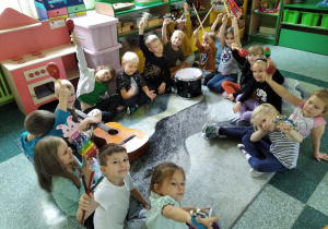 Dzieci z grupy Motylki siedzą na dywanie uśmiechnięte i trzymają w rękach instrumenty i machają nimi do zdjęcia. Na dywanie leży prawdziwa gitara.