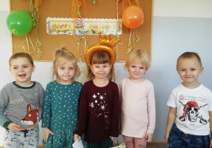 Po środku grupki dzieci stoi dziewczynka z koroną na głowie, w tle napis Happy Birthday.