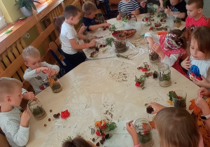 Dzieci zgromadzone są wokół dwóch złączonych stolików na których rozłożone są wszystkie potrzebne materiały do wykonania pracy (słoiki, piach, mech, liście, szyszki, kasztany, żołędzie). Każde dziecko dekoruje swój słoik według własnego pomysłu.