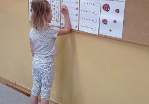 Dziewczynka ubrana w stój w kropki stoi przodem do tablicy na której przypięty jest napis:” Dzień Kropki” oraz karty pracy. Dziewczynka rozwiązuje zadania z trzeciej karty od lewej strony.