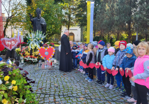 Dzieci stoją przed pomnikiem Jana Pawła II i rozmawiają z księdzem.