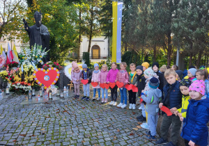Przedszkolaki stoją w rzędzie przy pomniku Jana Pawła II, trzymają w ręku czerwone serduszka.
