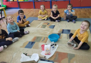 Dzieci z grupy Stokrotki siedzą w kole na dywanie a w środku znajdują się składniki oraz przedmioty, dzięki którym będą mogły przygotować chleb.