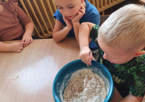 Chłopiec z grupy Słoneczka w zielonej podkoszulce miesza w misce mąkę z wodą. Obok obserwuje go chłopiec w niebieskiej koszulce.