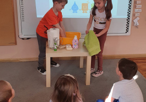 Dzieci z grupy Smerfy stoją przed tablicą interaktywną, na której wyświetla się napis „Światowy Dzień Mycia Rąk”. Dzieci pokazują reszcie grupy, jak prawidłowo myć ręce.