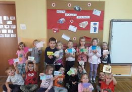 Dzieci z grupy „Żabki” pozują do zdjęcia na tle dekoracji z okazji Dnia Listonosza. Dzieci trzymają w ręku wykonane przez siebie prace plastyczne.