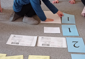 Chłopiec w niebieskiej bluzce z grupy „Pszczółki” siedzi na dywanie, układa kartonik z cyfrą „1” przy jednej kopercie. Na dywanie posegregowane są również inne druki pocztowe przy których ułożone są kartoniki odpowiadające liczbie druków.