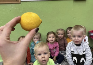 Nauczycielka prezentuje dzieciom przyniesione warzywa i owoce. Pokazuje cytrynę, a dzieci patrzą zaciekawione i odgadują co to za owoc.