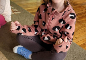 Uśmiechnięta dziewczynka siedzi na dywanie z nóżkami na kokardkę. Przed nią ułożony jest emblemat jeża z cyferką 5 i 5 emblematów dżdżownic.