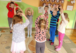 Dziewczynki i chłopcy w zabawie ruchowej stoją na dywanie z uniesionymi nad głowami rączkami.
