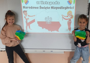 Dwie dziewczynki prezentują wykonaną przez siebie pracę plastyczną. Jest to mapa Polski. W tle tablica, na której jest napis 11 listopada Narodowe Święto Niepodległości.