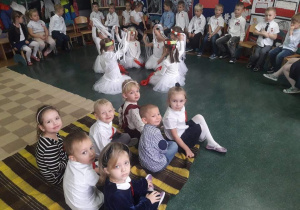 Przedszkolaki siedzą na kocu i oglądają taniec innych przedszkolaków.