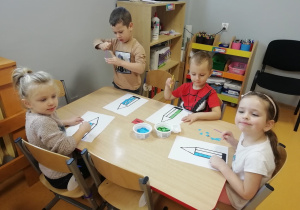 Dzieci siedzą przy stoliku i wyklejają kontur kredki kolorowym papierem.