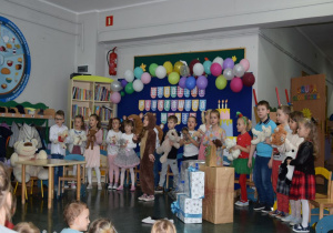 Dzieci występują przed publicznością, ustawione w rządku przed dekoracją z okazji Dnia Pluszowego Misia. W rękach trzymają swojego ulubionego pluszaka. Przed dziećmi stoją prezenty przyniesione dla misia.