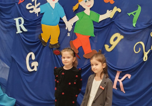 Dwie dziewczynki stoją na tle dekoracji przygotowanej na konkurs logopedyczny.