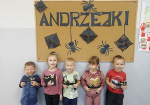 Dzieci trzymają w rękach papierowe koty i stoją na tle tablicy z napisem „Andrzejki”.