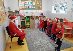 Na krześle siedzi Mikołaj, a przed nim stoją przedszkolaki.