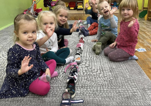 Dzieci machają do zdjęcia siedząc na dywanie przy ułożonych swoich bucikach jeden za drugim. Za chwilę dowiedzą się, kto będzie miał powodzenie.