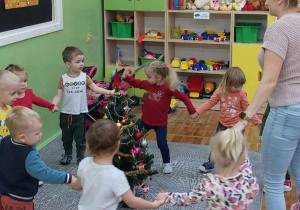 Dzieci tańczą na dywanie wokół pięknie ustrojonego świątecznego drzewka.