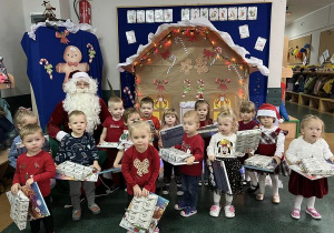 Dzieci uśmiechnięte pozują na tle dekoracji z okazji Mikołajek. W rękach trzymają prezenty, wraz z nimi do zdjęcia pozuje Mikołaj.
