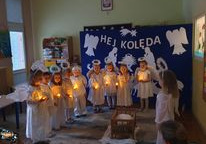 Dzieci przebrane za aniołki stoją na dywanie w rączkach trzymają światełka. Na środku stoi żłóbek. W tle widać dekoracje z napisem hej Kolęda