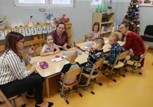 Dzieci i rodzice siedzą dookoła stolików a nauczycielka pokazuje karteczkę.