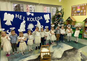 Zdjęcie grupowe dzieci przebranych za aniołki na tle niebieskiej dekoracji. Dzieci stoją w szeregu, a przed nimi na dywanie Jezusek w kołysce na sianku.