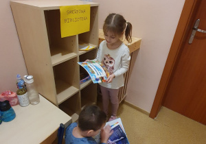 Na zdjęciu widać chłopca i dziewczynkę zastanawiających się nad wyborem książki. Biblioteką jest szafka z napisem „Smerfowa Biblioteka”, w której ułożone są książki. Nad szafką wypisane są zasady korzystania z książek.