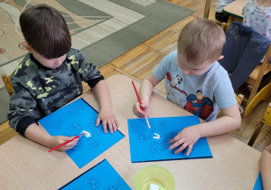 Dwaj chłopcy siedzą przy stoliku i malują białą farbą, niebieskie szablony śnieżynek