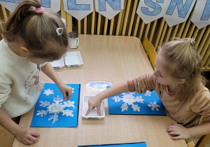 Dwie dziewczynki siedzą przy stoliku i posypują solą swoje pomalowane farbą śnieżynki