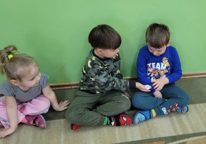 Dziewczynka z dwoma chłopcami siedzą na dywanie, chłopcy podają sobie guzik