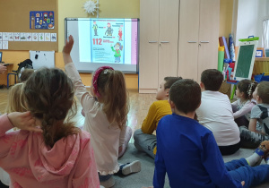 Dzieci siedzą na dywanie wpatrzone w prezentację, na której widoczne są nr alarmowe. Dziewczynka zgłasza się do odpowiedzi na pytanie: W jaki sposób reagujemy gdy komuś dzieje się krzywda?