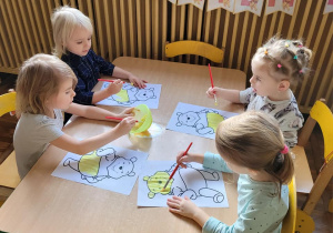 Cztery dziewczynki siedzą przy stoliku i malują żółtą farbą szablon Kubusia Puchatka