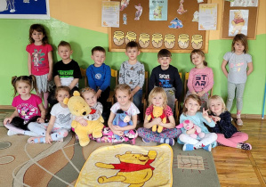 Wspólne zdjęcie dzieci z przyniesionymi maskotkami Kubusia Puchatka i jego przyjaciół na tle tablicy z Kubusiową dekoracją. Przed dziećmi rozłożony jest kocyk z wizerunkiem Kubusia Puchatka