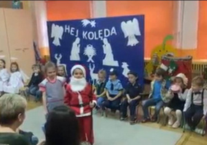 Dziewczynka w przebraniu jako dozorczyni stoi obok św. Mikołaja natomiast reszta dzieci siedzi.
