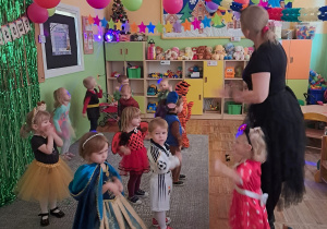 Dzieci tańczą wesoło do muzyki razem z Panią. Dookoła wiszą kolorowe dekoracje, girlandy, balony. W tle Sali świeci dyskotekowe światło.