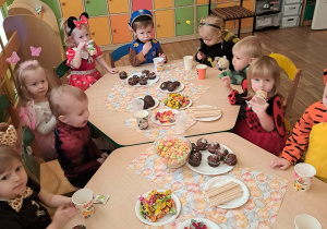 Dzieci siedzą dookoła stołu i zajadają pyszne smakołyki przygotowane przez rodziców. Widać pieczone babeczki, chrupki, ciasteczka, żelki i cukierki.