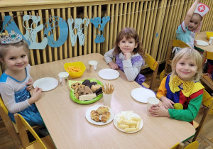 Na zdjęciu trzy dziewczynki w strojach Elzy, księżniczki i pajaca siedzą przy stoliku i jedzą słodycze.