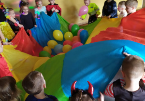Na zdjęciu dzieci z grupy Pszczółki stoją w kole, trzymając chustę na której leżą kolorowe balony.