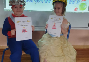 Chłopiec i dziewczynka siedzą na krzesełkach na tle tablicy z napisem bal karnawałowy. W koronach na głowach dumnie prezentują dyplomy Króla i Królowej Balu.