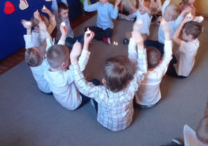 Dzieci prezentują występ muzyczno-ruchowy, chłopcy siedzą w kole z uniesionymi w górę lampeczkami a dziewczynki trzymają instrumenty muzyczne siedząc po bokach.