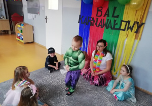 Dzieci siedzą z nauczycielka na dywanie pośrodku koła stoi chłopiec w stroju Hulka i trzyma w ręku chusteczka higieniczna.
