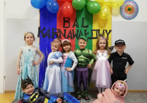Grupka przedszkolaków w strojach karnawałowych stoi pozuje do zdjęcia na tle kolorowej tablicy i napisu Bal Karnawałowy.