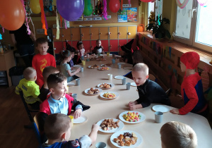 Dzieci siedzą przy stole na którym znajduje się słodki poczęstunek: kolorowe chrupki, chipsy, soki, cukierki itp.