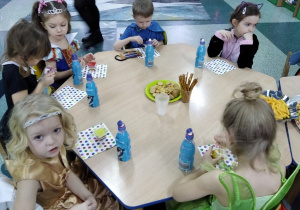 Dzieci siedzą przy stoliku i częstują się słodyczami przygotowanymi przez rodziców.