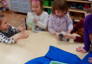 Dzieci siedzą przy stoliku, dwie dziewczynki przekazują sobie materiały, dotykają je i porównują ze sobą.